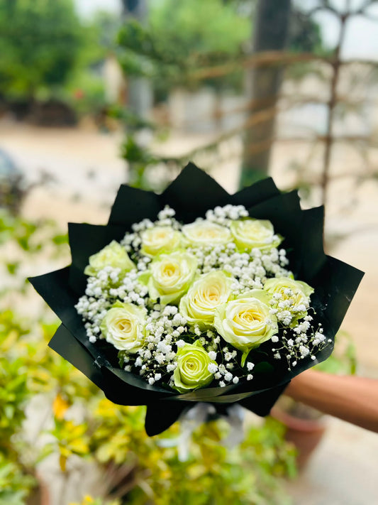 A bouquet of pale yellow roses and white baby's breath wrapped in black paper, held by a person's hand against a blurred outdoor garden background.